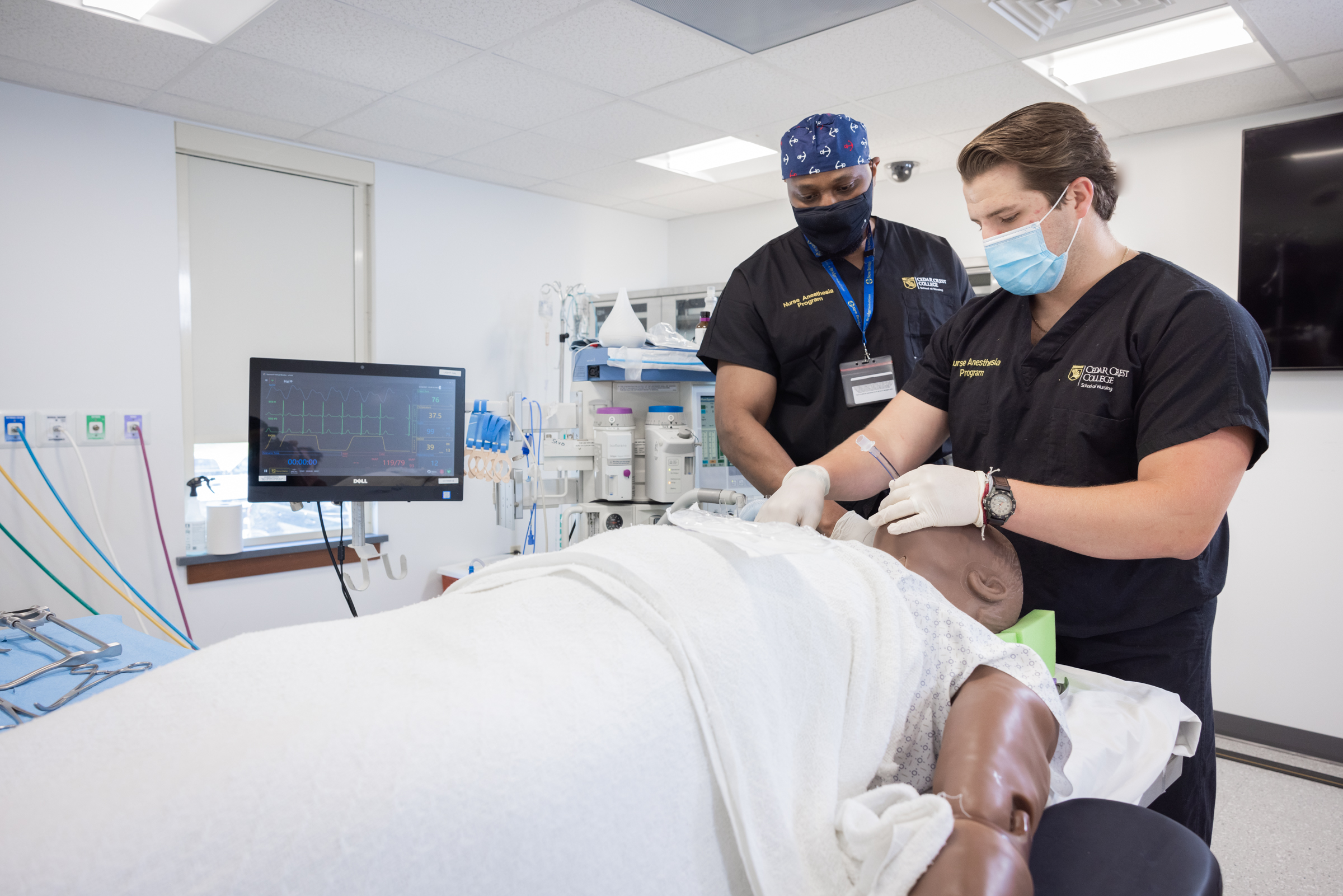 Two nurse anesthesia students wear masks as they tend to a manekin patient